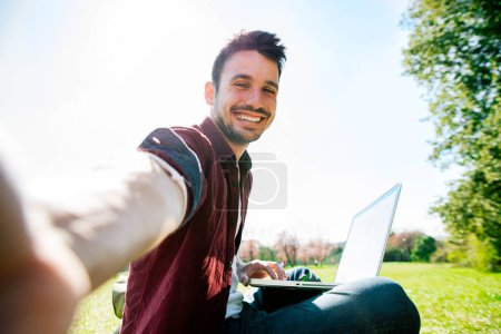 Photo for Happy male blogger taking a selfie in the park while working on laptop device using wireless internet - Remote work and social distance - Royalty Free Image