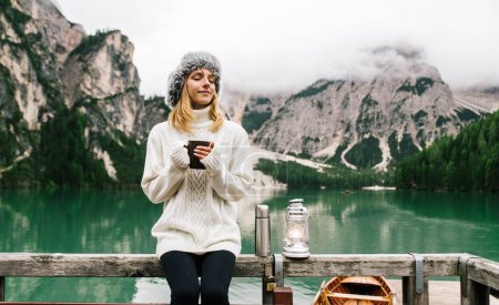 Foto de Retrato de una hermosa joven bebiendo sosteniendo taza de café caliente visitando un lago alpino en Braies Italia. Turista femenina disfrutando momentos de relax en las montañas. Personas, vagabundos y viajes. - Imagen libre de derechos