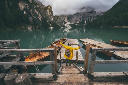 Téléchargez les photos : Vue arrière portrait d'un randonneur aux bras tendus visitant un lac alpin à Braies, en Italie. Joyeux randonneur vêtu d'une veste jaune et sac à dos profiter du paysage naturel à l'automne. Désir et voyage. - en image libre de droit