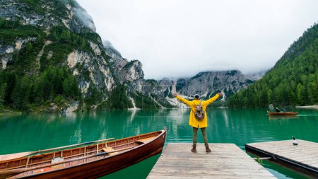 Téléchargez les photos : Vue arrière portrait d'un randonneur aux bras tendus visitant un lac alpin à Braies, en Italie. Joyeux randonneur vêtu d'une veste jaune et sac à dos profiter du paysage naturel à l'automne. Désir et voyage. - en image libre de droit