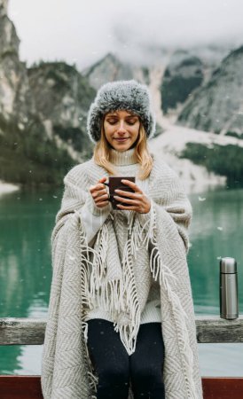 Photo for Portrait of a beautiful young woman drinking holding hot coffee cup visiting an alpine lake at Braies Italy. Female tourist enjoying moments relaxing at mountains. People, wanderlust and travel - Royalty Free Image