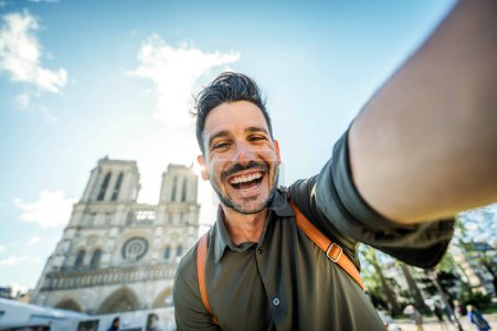 Téléchargez les photos : Un bel homme prend un selfie avec un téléphone portable intelligent dans la rue de la ville - Bonne visite touristique Notre Dame à Paris, France - Vacances d'été et concept d'influenceur de voyage - en image libre de droit