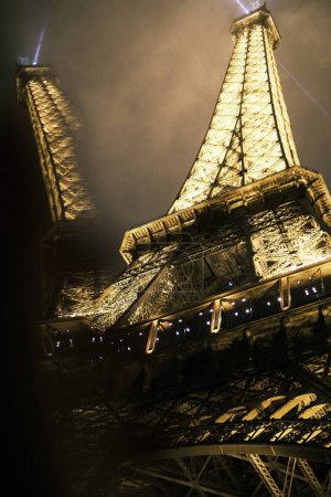 Téléchargez les photos : As night descends on Paris, the Eiffel Tower stands radiant, its lights a dazzling display against the nocturnal backdrop. This image portrays the tower's powerful beams cutting through the darkness, a testament to the city's enduring allure and the - en image libre de droit