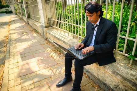 Businessman working on laptop outdoors, While sitting on a stone ledge, He is near metal fence with green plants background, A young business tycoon work with focus, Sitting on street pathway, Indian.