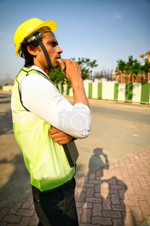 Photo for Thoughtful construction worker at a site, Wearing a yellow hard hat reflective vest, Standing outdoors, Looking thoughtful, He is holding a tablet, And has his hand on his chin, With barriers, Indian. - Royalty Free Image