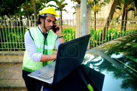 Construction engineer working on laptop and talking on phone, wearing a yellow hard hat and reflective vest, is talk on mobile, While work on a computer, Placed on the boot of a car, fence, 30s guy.