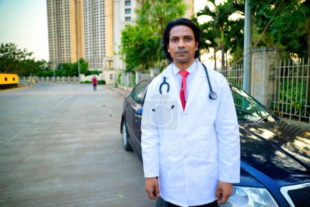 Männlicher Arzt, der im Freien neben einem geparkten Auto steht, Arzt mit weißem Mantel und roter Krawatte, Steht draußen, Gebäude und Bäume, Hintergrund, Auf der Stadtstraße, Inder, Junger Chirurg blickt in die Kamera.