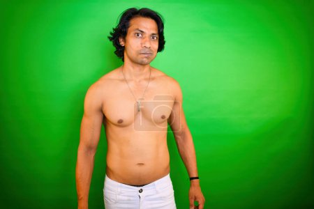Hemdloser Mann mit ausdrucksstarkem Blick, steht vor grünem Hintergrund, trägt weiße Hosen und eine Halskette, er hat ein beredtes Gesicht, und seine Arme sind ausgestreckt, indisch, asiatisch.