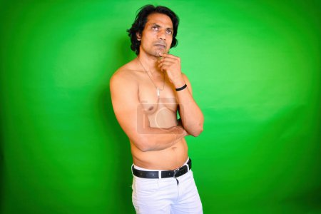 Téléchargez les photos : Shirtless man posing thoughtfully, Men thinking pose, Looking up while think, Indian, With long hair, stands against a green background, Looks thoughtful, Hand on his chin, White pant, Pensive male. - en image libre de droit