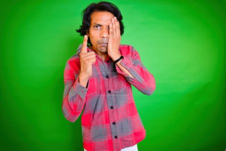 Téléchargez les photos : Man covering one eye and threatening, Person threat someone, Angry man got slapped, checkered shirt stands against a green background, While raising his index finger, Indian, Slap Hit his face, 30s. - en image libre de droit