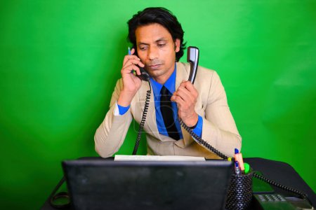 Geschäftsmann Gespräch auf zwei Telefonen, Geschäftsmann Multitasking mit Telefon und Dokumenten, Ein Mann jongliert am Schreibtisch, Während er auf Laptop schaut, trägt einen beigen Anzug, grünen Hintergrund, Indianer, Jonglieren.