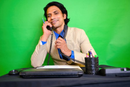Smiling businessman multitasking with two phones, A business person in beige suit, Talking on landlines simultaneously, While sitting at a desk, Green background, Indian, Happy entrepreneur at office.