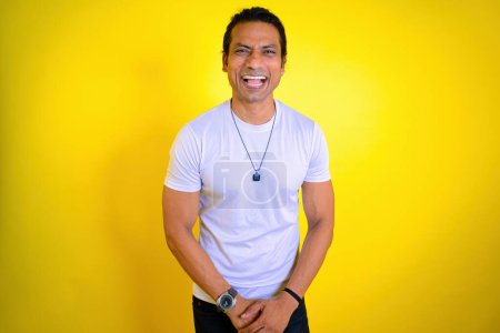 Man in white t shirt laughing against a yellow background. Laughing madly in studio. expressing happiness. toothy smile. Dental care. Indian. Laughter dental concept. Man enjoying success, Asian, 30s