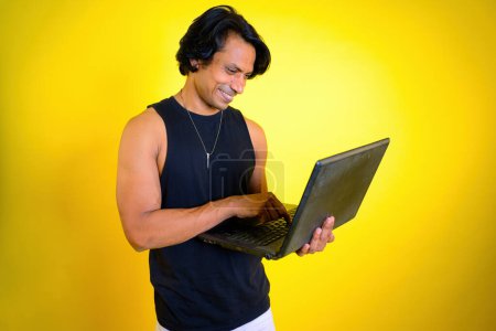 Mann mit Laptop vor gelbem Hintergrund, Lächeln fokussiert Man Working On, Person in schwarzem Tanktop, Office-Arbeitszeit, Computer in der Hand, Tippen auf der Tastatur, Video-Chat-Sitzung, indisch, asiatisch, 30er Jahre,