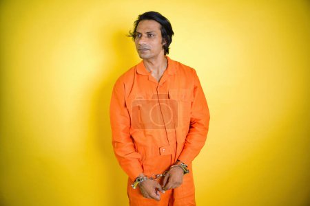 Smiling prisoner standing with shackles, Man in an orange jumpsuit handcuffed, posing against a yellow background, Looking away, Indian, Studio, fetters chained, slight smile, 30s prison, long hairs