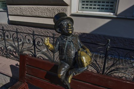 Téléchargez les photos : Sculpture d'un gai gnome coiffé d'un chapeau haut de gamme.Petite figurine d'un homme assis sur la tête d'un banc de rue. Attire l'attention des touristes. - en image libre de droit