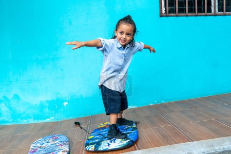 Photo for A cute boy with long hair practicing surfing in different poses at home.stand on the surfboard. wearing clothes gracefully.cheerful smiley face. blue wall background. - Royalty Free Image