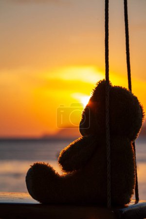 Téléchargez les photos : Lonely Teddy Bear assis sur une balançoire en face de la plage de Rawai, reflet pittoresque de beau soleil au-dessus des bateaux de pêche. - en image libre de droit