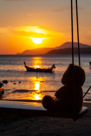 Foto de Lonely Teddy Bear sentado en un columpio frente a la playa de Rawai, reflejo escénico del hermoso sol sobre los barcos de pesca. - Imagen libre de derechos
