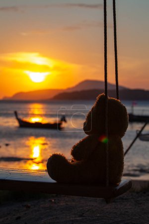 Photo for Lonely Teddy Bear sitting on a swing in front Rawai beach, scenic reflection of beautiful sunshine above fishing boats. - Royalty Free Image