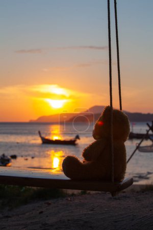 Téléchargez les photos : Lonely Teddy Bear assis sur une balançoire en face de la plage de Rawai, reflet pittoresque de beau soleil au-dessus des bateaux de pêche. - en image libre de droit