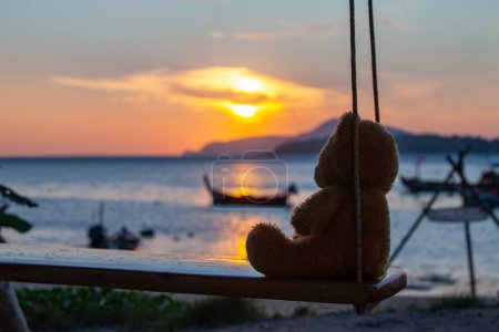 Foto de Lonely Teddy Bear sentado en un columpio frente a la playa de Rawai, reflejo escénico del hermoso sol sobre los barcos de pesca. - Imagen libre de derechos
