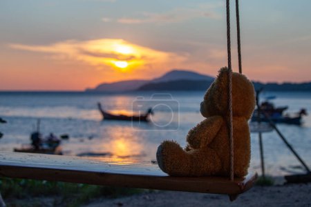 Photo for Lonely Teddy Bear sitting on a swing in front Rawai beach, scenic reflection of beautiful sunshine above fishing boats. - Royalty Free Image