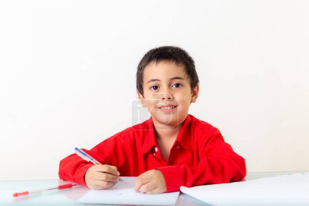 Foto de Un chico lindo con camisa roja sentado y escribiendo, haciendo los deberes. Estudio retrato, concepto de salud con fondo blanco. - Imagen libre de derechos