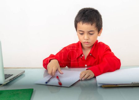 Photo for A cute boy in red shirt sitting and writing, doing homework. Studio portrait, concept health with white background. - Royalty Free Image