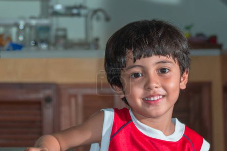 Téléchargez les photos : Asiatique petit garçon portant rouge t-shirt assis dans la cuisine en attendant un repas - en image libre de droit