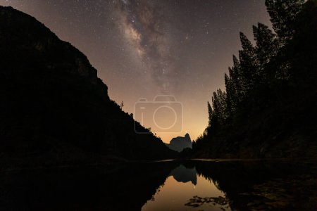 Photo for It is a stunning sight as the Milky Way towers over a large pond in a quarry.On one side is a pine forest and on the other is a mountain. The reflection of the stars in the water at night - Royalty Free Image