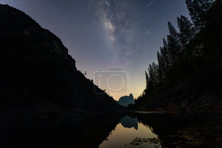 Photo for It is a stunning sight as the Milky Way towers over a large pond in a quarry.On one side is a pine forest and on the other is a mountain. The reflection of the stars in the water at night - Royalty Free Image