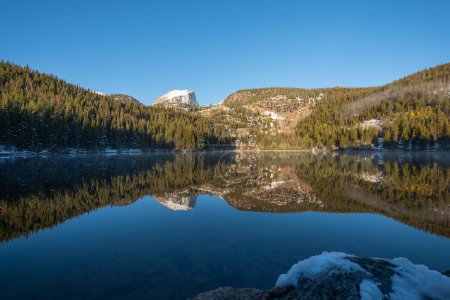 Téléchargez les photos : Paysage d'un lac alpin avec un reflet miroir au coucher du soleil. Un pic enneigé s'élève au-dessus de la forêt dans le ciel, et de la neige légère est visible à la base des arbres et sur un rocher au premier plan. - en image libre de droit