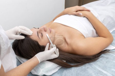 junge Frau liegt auf einer Trage in einem ästhetischen Zentrum und führt Gesichts- und Körperschönheiten sowie ästhetische Behandlungen mit Dermapen und Dermaplaning-Techniken mit Skalpell durch