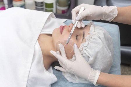 junge Frau liegt auf einer Trage in einem ästhetischen Zentrum und führt Gesichtsschönheit und ästhetische Behandlung mit Dermaplaning-Techniken und Skalpell durch