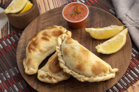 Téléchargez les photos : Empanadas maison cuites avec des légumes de viande de poulet et jambon et fromage sur une assiette en bois avec des sauces et du citron - en image libre de droit