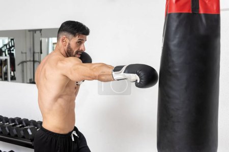 Téléchargez les photos : Homme musclé dans la salle de gym pratiquant la boxe et l'entraînement avec un sac et des gants de boxe - en image libre de droit