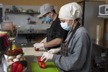 Téléchargez les photos : Deux jeunes entrepreneurs cuisinant dans une cuisine professionnelle avec gants et masque pour les soins d'hygiène alimentaire - en image libre de droit