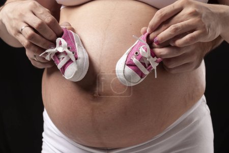 Téléchargez les photos : Femme enceinte montrant son ventre avec une paire de petites baskets avec ses mains sur son ventre et celles de son père - en image libre de droit