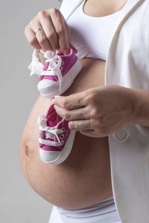 Téléchargez les photos : Femme enceinte montrant son ventre avec une paire de petites baskets avec ses mains sur son ventre et celles de son père - en image libre de droit