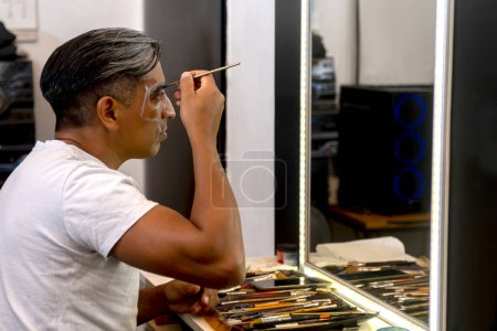 Téléchargez les photos : Homme se maquillant devant un miroir dans son atelier de peinture et de maquillage peignant son visage pour la célébration des morts - en image libre de droit