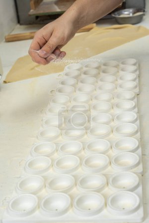Téléchargez les photos : Fabrication de pâtes maison mains remplissant les sorrentinos et étirant la pâte dans des moules avec le remplissage dans une cuisine - en image libre de droit
