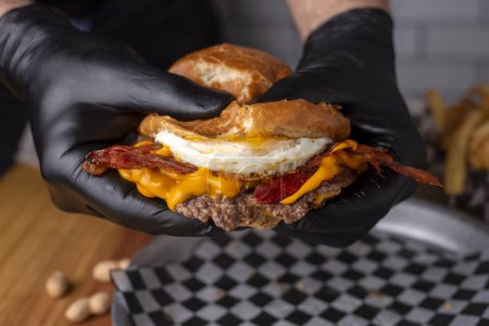 Téléchargez les photos : Mains tenant un grand hamburger de viande au fromage jambon bacon oignon tomate pomme de terre pain cheddar laitue frites - en image libre de droit