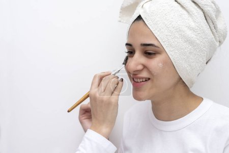 Téléchargez les photos : Jeune femme dans un centre de beauté avec un compte-gouttes à la main effectuant un traitement cosmétique sur son visage avec une chemise et une serviette sur sa tête et fond blanc - en image libre de droit