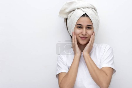 Téléchargez les photos : Jeune femme dans un centre de beauté avec un compte-gouttes à la main effectuant un traitement cosmétique sur son visage avec une chemise et une serviette sur sa tête et fond blanc - en image libre de droit