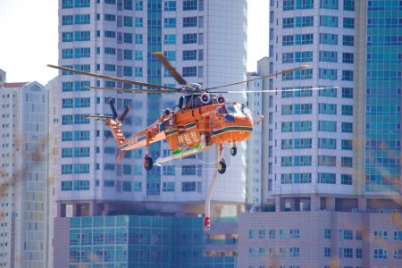 Foto de Un helicóptero de extinción de incendios desciende para aterrizar, enmarcado por imponentes edificios de apartamentos en el fondo, fusionando paisajes urbanos con aviación de emergencia. - Imagen libre de derechos