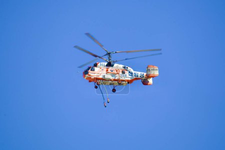 Téléchargez les photos : Capturé contre un ciel bleu clair, un hélicoptère de lutte contre les incendies vole avec des tuyaux suspendus en dessous, prêts pour une intervention d'urgence rapide. - en image libre de droit