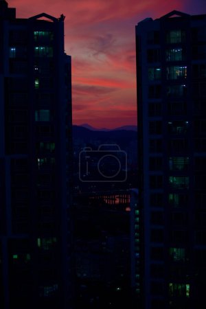 Foto de El horizonte de Ulsan al atardecer, con siluetas de apartamentos oscuros, cielo vibrante, río Taehwa sereno y el icónico puente Taehwa. - Imagen libre de derechos