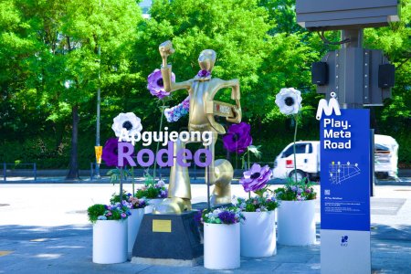 Foto de Seúl, Corea del Sur - 2 de junio de 2023: El dinámico cartel de Apgujeong Rodeo con una escultura de hombre dorado en un traje, rodeado de flores rosas y púrpuras, encarna la elegante esencia de la zona. - Imagen libre de derechos