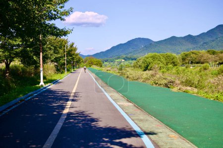 Photo for Hanam City, South Korea - October 1, 2023: A serene bike path lined with trees, adjacent to a walking path, with a cyclist enjoying a ride beside the scenic riverside mountains. - Royalty Free Image